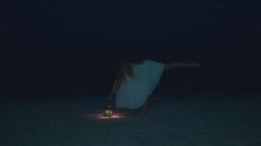 Beyaz giyinmiş bir balerin gibi hareket eden bir kadın, gece kumsalda fener tutuyor, karanlık gökyüzü ve kumla çakışıyor.