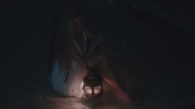 Huzurlu bir gecede, beyaz elbiseli bir kadın elinde fenerle okyanusun kenarında oturur, yalnızlığı ve huzuru temsil eder.