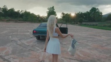 Gün batımında sakin bir açık havada, güzel sarışın bir kadın yeşil bir arabaya doğru yürür.