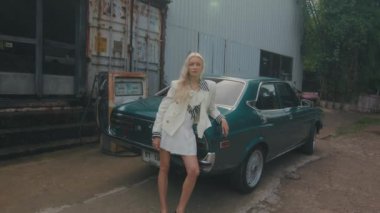 Moda düşkünü beyaz elbiseli genç bir kadın endüstriyel bir yerin yakınında klasik yeşil bir arabanın yanında duruyor..