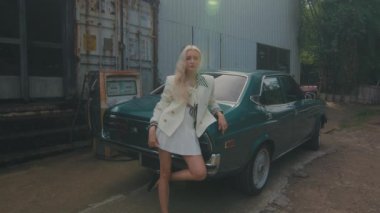 Beyaz elbiseli ve ceketli sarışın kadın sanayi bölgesinde klasik arabanın yanında güven içinde poz veriyor..