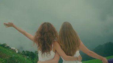 Beyaz elbiseli iki genç kız sisli, rüya gibi bir atmosferde güzel çiçekler arasında hassas bir anı paylaşıyor.