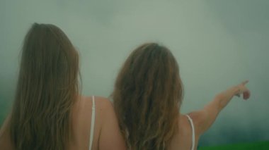 Beyaz elbiseli iki genç kız sisli, rüya gibi bir atmosferde güzel çiçekler arasında hassas bir anı paylaşıyor.