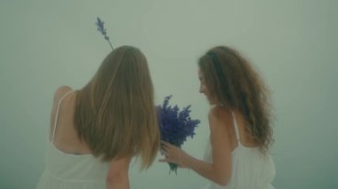 Beyaz elbiseli iki genç kız sisli, rüya gibi bir atmosferde güzel çiçekler arasında hassas bir anı paylaşıyor.