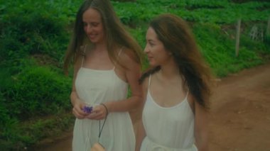 Beyaz elbiseli iki kadın neşe içinde güzel yemyeşil bir yolda yürüyorlar, birlikte doğanın tadını çıkarıyorlar.