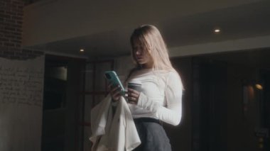 Genç bir kadın şık bir kapalı mekan ortamında zarif bir şekilde duruyor, akıllı telefon cihazına derinden odaklanmış.