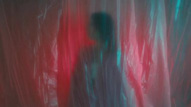 Pembe ceketli genç bir kadın plastik filmin arkasında neon ışıkta hareket ediyor.