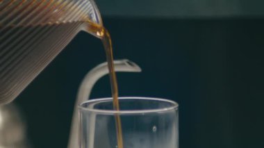 Temiz bir bardağa dökülen aromatik kahvenin yakın çekimi, karışık demleme sürecini gözler önüne seriyor.
