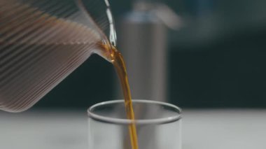 Temiz bir bardağa dökülen aromatik kahvenin yakın çekimi, karışık demleme sürecini gözler önüne seriyor.