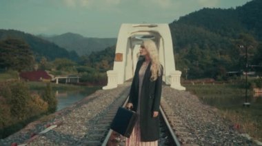 Huzurlu bir sahnede, görkemli dağların ve yemyeşil yeşilliklerin ortasındaki pitoresk bir demiryolu köprüsünde eski moda kıyafetler içindeki bir kadın görülüyor.