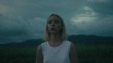 Düşünceli ve düşünceli bir kadın dramatik ve karanlık bulutların altında engin, açık bir alanda zarif bir şekilde duruyor.