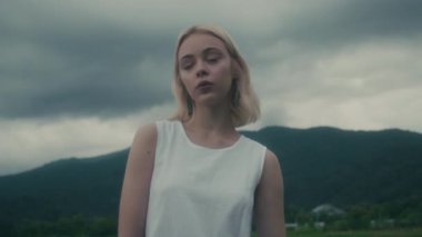 Uçsuz bucaksız bulutlu gökyüzünün altındaki yemyeşil bir tarlada dalgalı beyaz elbiseli bir kadın duruyor.