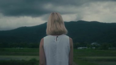 Uçsuz bucaksız, bulutlu gökyüzünün altındaki yemyeşil bir tarlada zarif bir şekilde yürüyen beyaz elbiseli bir kadın.