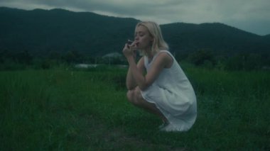 Genç bir kadın yeşil yemyeşil bir tarlada düşünceli bir şekilde oturuyor, elinde sigarayla, bir an yansımasını yakalıyor..