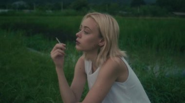 Genç bir kadın yeşil yemyeşil bir tarlada düşünceli bir şekilde oturuyor, elinde sigarayla, bir an yansımasını yakalıyor..