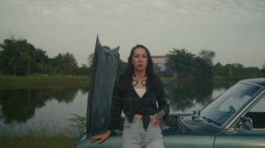 Taşralı bir kadın, bir göl kenarındaki köy yolunda bozulmuş bir eski arabanın yanında duruyor.