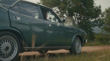 Bir kadın, kaputun altından çıkan dumanı gördüğünde aniden kırsal bir yolda bozulan eski bir arabadan iner. 