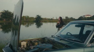 Bir kadın ne yapacağını bilmez ve arka planda bir gölün önünde duran eski bir arabanın yanından geçer.