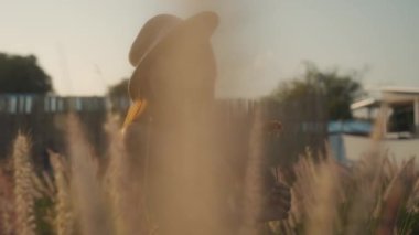 Yeşil yazlık elbiseli genç bir kadın, sık otların içinde çayırlarda yürüyor ve eli, güneşli bir günde yavaş çekimde çiçeklerin tepelerine değiyor. Yüksek kalite 4k görüntü