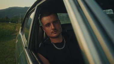 Kendine güvenen genç bir adam, nefes kesen doğal bir arka planla çevrili klasik bir arabada poz veriyor.