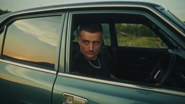 Düşünceli genç bir adam eski bir arabada oturup nefes kesici güneşin batışının tadını çıkarıyor.