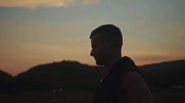 Genç bir adam çarpıcı bir siluette duruyor, düşünceli bir şekilde sigara içiyor ve canlı bir gün batımına bakıyor.