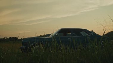 Gün batımında manzaralı bir araziye park edilmiş klasik bir araba. Etrafı dağlar ve doğal güzelliklerle çevrili..