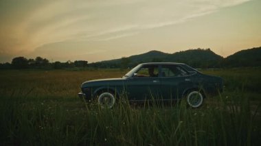 Gün batımında manzaralı bir araziye park edilmiş klasik bir araba. Etrafı dağlar ve doğal güzelliklerle çevrili..