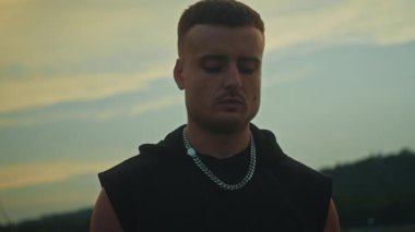 Genç bir adam çarpıcı bir siluette duruyor, düşünceli bir şekilde sigara içiyor ve canlı bir gün batımına bakıyor.