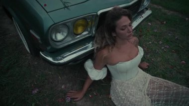 Çarpıcı beyaz elbiseli bir kadın klasik bir arabanın yanında oturmuş, doğanın güzelliğinin ortasında bir anı yakalıyor.