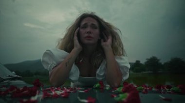 Son derece dokunaklı bir sahne, kasvetli ve bulutlu bir gökyüzünün altında canlı çiçek yapraklarıyla çevrili sıkıntılı bir kadını sergiliyor.