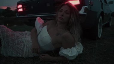 Çarpıcı bir sahnede beyaz elbiseli bir kadın gün batımında klasik bir arabanın yanında yatıyor, derin duygular uyandırıyor.