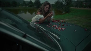 Huzurlu ve huzurlu bir anda dağınık çiçek yaprakları arasında eski bir arabanın kaputunda dinlenen bir kadın görülüyor.