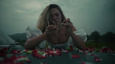 Yumuşak, bulutlu bir günde, etrafını narin çiçek yapraklarıyla çevirmiş bir kadın duygularını derinden ifade eder.