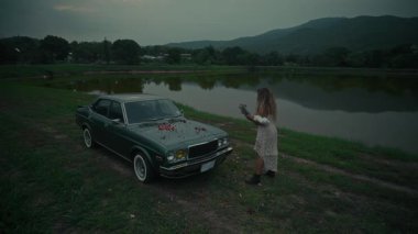 Renkli çiçeklerle bezenmiş eski bir arabanın yanında duran bir kadın, yakınlardaki dingin gölün yanında duyguları uyandırıyor.