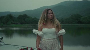 Huzurlu bir kadın durgun suyun yanında zarif bir şekilde durur, rüya gibi bir atmosferi çağrıştıran dalgalı bir elbiseyle süslenir.