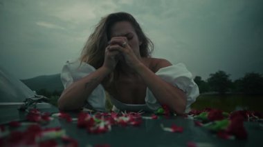 Yumuşak, bulutlu bir günde, etrafını narin çiçek yapraklarıyla çevirmiş bir kadın duygularını derinden ifade eder.