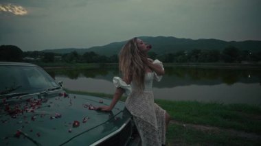 Dalgalı beyaz elbiseli zarif bir kadın gölün kenarında poz veriyor ve renkli çiçek yapraklarıyla süslenmiş klasik bir araba.