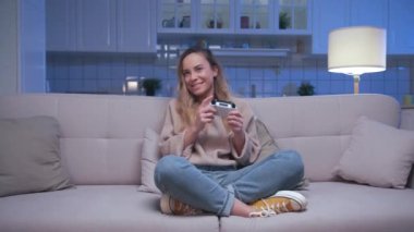 Koltukta oturan ve Dijital Konsolda Video Oyunlarında oynayan Mutlu Genç ve Güzel Oyuncu Kadın. Video oyunu için Joy Reaction Player Girl Joystick 'i kontrol ediyor. Oturma odasında oyun oynamaya odaklanmış biri.