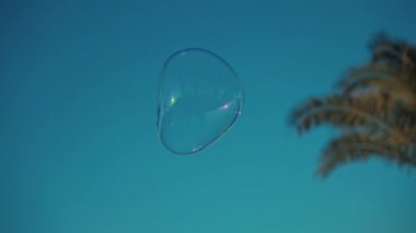 Bir sabun köpüğü mavi renkli gökyüzünde uçuyor. Köpük Fırtınası ile Mutlu Oyuncu Yaz Gününde dışarıda eğleniyor. Soapy Water 'dan Kabarcıklı Yuvarlak Yüzen Top Şekliyle Açık Hava Oyunu