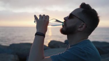 Genç Yetişkin Kişi Dışarıda Güneş Işığında Dinleniyor ve İçiyor. Cazip Kafkasyalı Adam Ocean Close 'un Akşam Güneşinde İçecek İçiyor. Bahar Gökyüzünde Modern Aktif Eğlence ve Sahne Güneşi