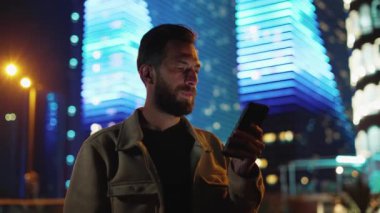 Urban Caddesi 'nde elinde telefon olan bir adam internette arama yapıyor. Neon Dark Evening 'de Yürüyen ve Fütüristik Aydınlatılmış Binalara Yakın Akıllı Telefon Kullanıyor. İnsanların Büyük Yaşamı İçin Elektronik Bilgi