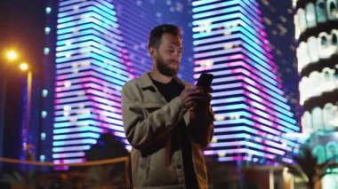 Bir Sakallı Adam Karanlık Şehir Caddesinde Haberleri okuyor. Neon Evening Binası 'nın dışında Smartphone' lu biri var. Touchscreen 'de Sosyal Medyayla Elektronik Görüntüyle Turist Yürüyüşleri