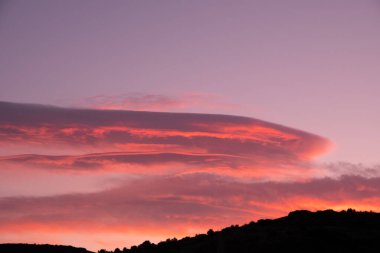 Bir kaç kümülüs bulutu olan pitoresk bir günbatımı gökyüzü, turuncu ve kırmızı tonlarıyla çarpıcı bir atmosfer yaratıyor. Doğal manzara alacakaranlıktan sonra yıkanır.