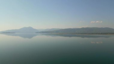 Arka planda sisli dağlarla çevrili, gölü yansıtan sakin doğal bir manzara..