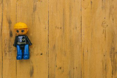 Sarı kasklı, mavi çizmeli, ahşap arka planda gülümseyen elsiz lego işçisi.