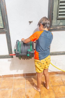 Genç bir çocuk yeşil bahçe hortumunu makaraya sarmaya odaklanmıştı..