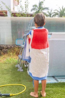 Kırmızı ve mavi çizgili havluya sarılmış bir çocuk düşünceli bir bakışla havuzun kenarında duruyor..