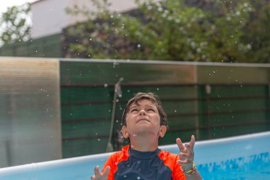 Sıcak bir yaz gününde yüzme havuzunda baş parmak gösteren turuncu yüzme tişörtlü gülen çocuk.
