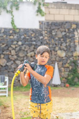 Islak spor elbiseli, bahçe hortumunu tutan, güneşli bir bahçede yüzünde odaklanmış ifadeyle genç bir çocuk..